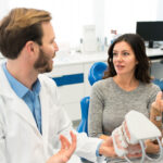 The image depicts a dental professional showing a dental model to a patient. The patient is attentively listening and engaged in the conversation.