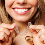 a woman smiling and holding up an invisalign clear aligner that highlights her straight teeth